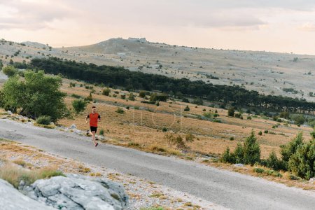 Ein muskulöser männlicher Athlet läuft bei Sonnenaufgang auf einem schroffen Bergpfad, umgeben von atemberaubenden Felslandschaften und natürlicher Schönheit.. 