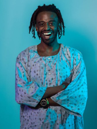 A Sudanese man adorned with modern dreadlocks stands proudly in traditional Sudanese attire, his arms crossed, conveying a blend of cultural heritage and contemporary style against a vibrant blue