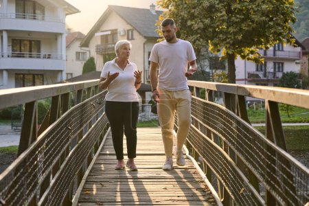 Ein gutaussehender Mann und eine ältere Frau teilen sich einen ruhigen Spaziergang in der Natur, überqueren eine schöne Brücke vor dem Hintergrund eines atemberaubenden Sonnenuntergangs und verkörpern das Konzept einer gesunden und dynamischen Generationenbeziehung