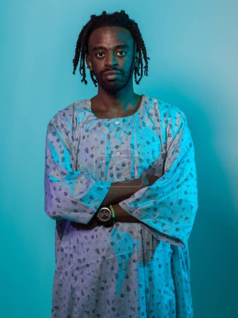 Ein sudanesischer Mann mit modernen Dreadlocks steht stolz in traditioneller sudanesischer Kleidung, die Arme verschränkt, und vermittelt eine Mischung aus kulturellem Erbe und zeitgenössischem Stil vor einem lebendigen Blau