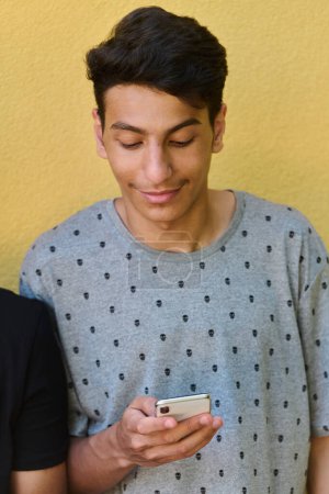  Middle Eastern teenager engrossed in his mobile phone while leaning against a vibrant yellow wall.
