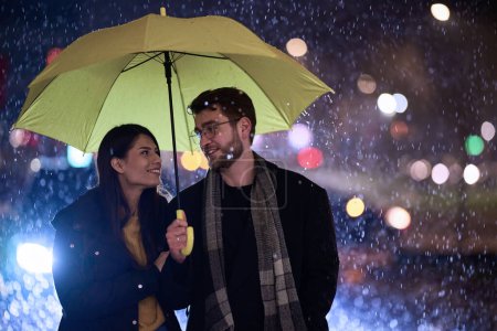 En el ambiente romántico de una noche lluviosa, una feliz pareja camina por la ciudad, compartiendo momentos tiernos bajo un paraguas amarillo, rodeado por el resplandor brillante de las luces urbanas.