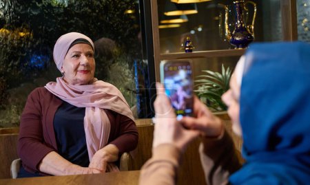 In einem modernen Restaurant während des heiligen Fastenmonats Ramadan hält eine Frau im Hidschab einen Moment mit ihrer Mutter mit einem Smartphone fest und verkörpert die Verschmelzung von Tradition und Technologie in familiären Bindungen