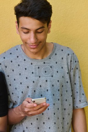  Adolescente de Oriente Medio absorto en su teléfono móvil mientras se apoya en una vibrante pared amarilla.