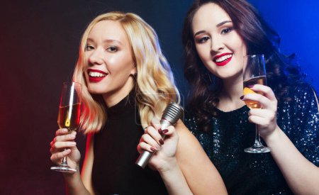 Téléchargez les photos : Célébration, fête et concept people - deux jeunes femmes chics en robes de cocktail noires chantent des chansons avec un microphone et tiennent des verres de champagne. - en image libre de droit