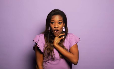 Téléchargez les photos : Jeune femme afro-américaine est étonnée avec la bouche ouverte, regarde la caméra dans la surprise. Portrait sur fond de couleur violette. - en image libre de droit