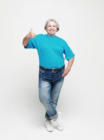 Téléchargez les photos : Mode de vie, émotion et concept humain : Femme âgée heureuse portant un jean et une chemise bleue donnant un pouce vers le haut et regardant la caméra. Portrait pleine longueur. - en image libre de droit