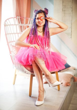 Foto de Mujer bonita joven caucásica con rastas peinado con vestido rosa, estilo muñeca, maquillaje brillante, hasta en la habitación rosa. Concepto del partido. - Imagen libre de derechos