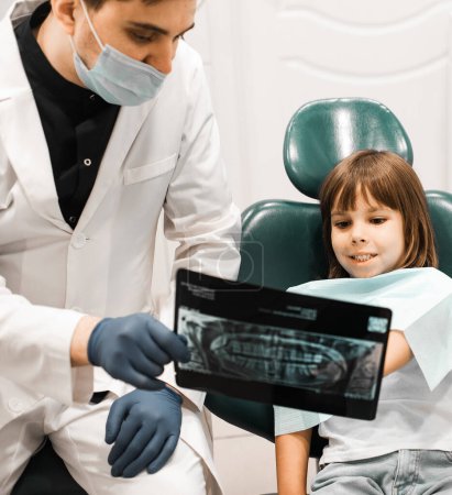 Foto de Odontólogo masculino mostrando radiografía de dientes a niña en consultorio dental. Personas, medicina, estomatología, tecnología y concepto de atención sanitaria. - Imagen libre de derechos