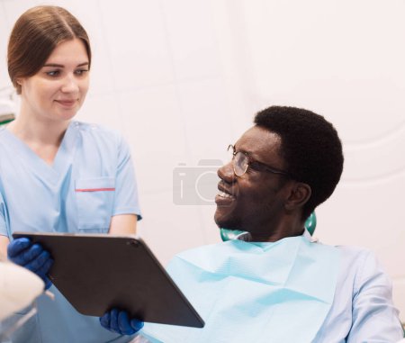 Photo for People, medicine, stomatology, technology and health care concept. Female dentist showing teeth x-ray to afro male at dental clinic office. - Royalty Free Image