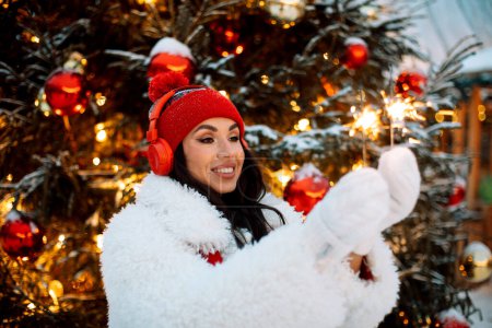 Photo for Charming young woman wearing red hat and headphones holding sparklers at the Christmas market next to the New Year tree. Happy time. - Royalty Free Image