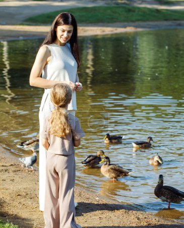 Foto de Mom and daughter feeding ducks in a park on the lake. Summer time. - Imagen libre de derechos