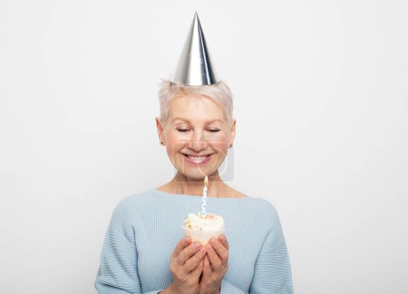 Téléchargez les photos : Beauitful elderly female wearing conical hat celebrating birthday with cupcake with candle over grey background - en image libre de droit