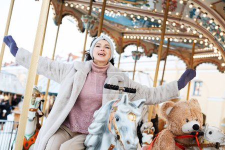 Photo for Beautiful and cheerful brunette woman in a fur coat rides on a carousel, Christmas winter fair. - Royalty Free Image