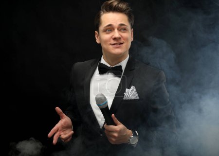 Photo for Stylish young man in a tuxedo holding a microphone, posing against a dark background with smoke, actor, singer, show, host of the event. Party concept. - Royalty Free Image