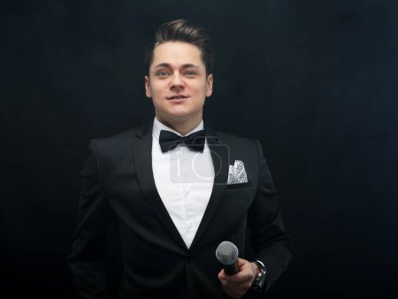 Téléchargez les photos : Stylish young man in a tuxedo holding a microphone, posing against a dark background with smoke, actor, singer, show, host of the event. Party concept. - en image libre de droit