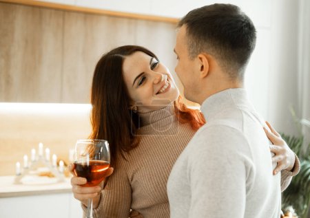 Foto de Una joven pareja enamorada abrazando y bebiendo vino tinto de copas en una cocina moderna. El concepto de amor y ternura. - Imagen libre de derechos