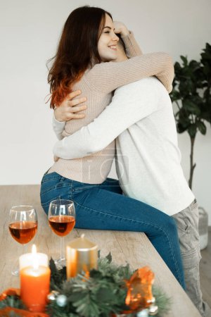 Foto de Una joven pareja enamorada abrazando y bebiendo vino tinto de copas en una cocina moderna. El concepto de amor y ternura. - Imagen libre de derechos