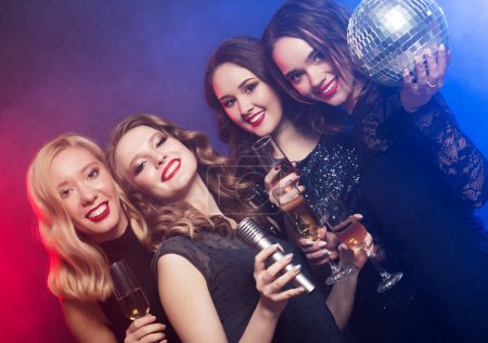 Photo for Celebration, party, and people concept - chic young women in black cocktail dresses sing songs with a microphone and hold glasses of champagne and disco balls. - Royalty Free Image
