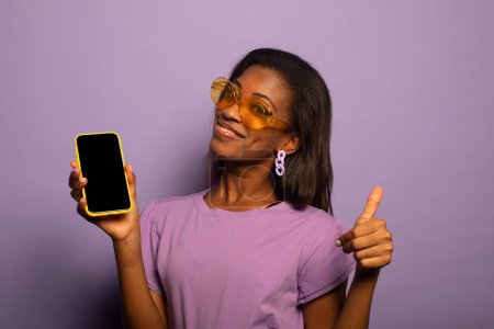 Photo for Portrait of positive young afro american woman promoter show smartphone, present new modern device wear lilac bluse isolated over violet color background - Royalty Free Image