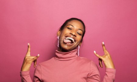 Photo for Young happy afro american woman in pink sweater smiling on camera and gesturing rock sign isolated over pink color background - Royalty Free Image