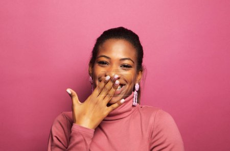 Foto de Joven mujer africana sorprendida sonriendo ante la cámara sobre fondo rosa. Joven hembra sostiene las manos cerca de su cara y sonríe ampliamente. Concepto de belleza. - Imagen libre de derechos