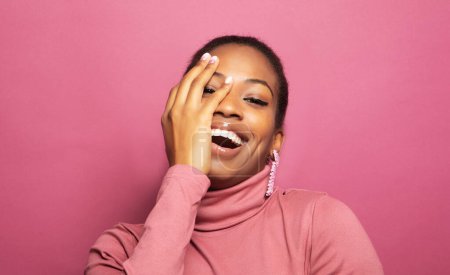 Foto de Joven mujer africana sorprendida sonriendo ante la cámara sobre fondo rosa. Joven hembra sostiene las manos cerca de su cara y sonríe ampliamente. Concepto de belleza. - Imagen libre de derechos
