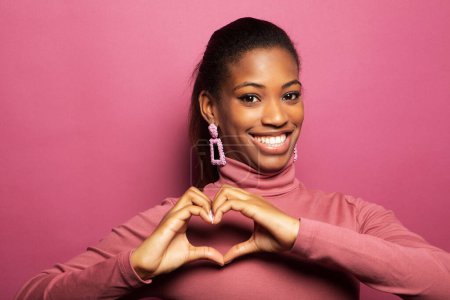 Foto de Una joven afroamericana se ríe alegremente y muestra un corazón de sus dedos. Retrato sobre fondo rosa. El concepto de amor, día de San Valentín. - Imagen libre de derechos