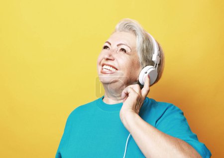 Téléchargez les photos : Portrait de femme âgée écoutant de la musique avec un casque sur fond jaune. - en image libre de droit