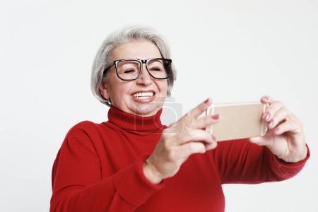 Téléchargez les photos : C'est l'heure du selfie. Image de bonne vieille femme mature debout isolé sur un mur de fond blanc parlant par téléphone mobile. - en image libre de droit