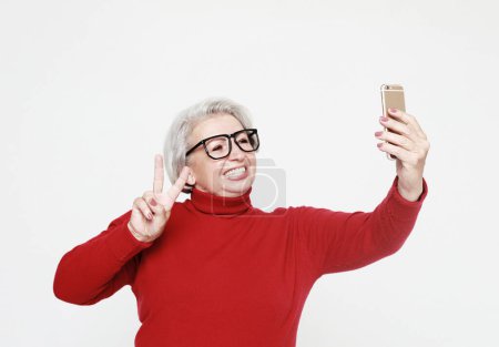 Téléchargez les photos : C'est l'heure du selfie. Image de bonne vieille femme mature debout isolé sur un mur de fond blanc parlant par téléphone mobile. - en image libre de droit