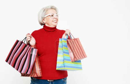 Foto de Foto de positiva alegre gris pelo blanco anciano mujer disfrutar del tiempo libre compras celebrar bolsas usar jeans y suéter rojo aislado sobre fondo blanco - Imagen libre de derechos