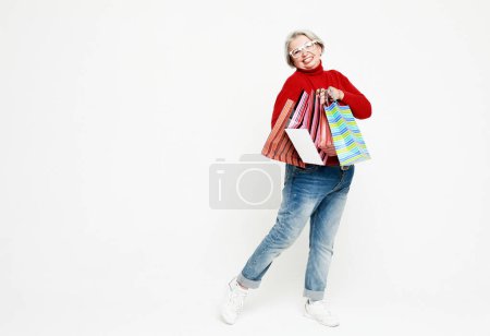 Foto de Foto de longitud completa de la mujer de edad avanzada positivo alegre pelo blanco gris disfrutar de tiempo libre bolsas de sujeción de compras usan jeans y suéter rojo aislado sobre fondo blanco - Imagen libre de derechos