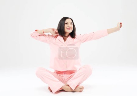 Téléchargez les photos : Jeune jolie femme asiatique en pyjama rose assise sur le sol et étirant, étirant, yoga, éveil. - en image libre de droit
