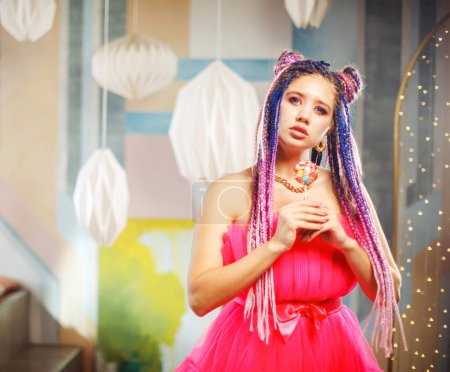 Foto de Mujer bonita joven caucásica con rastas peinado con vestido rosa, estilo muñeca, maquillaje brillante, hasta en la habitación rosa. Concepto del partido. - Imagen libre de derechos