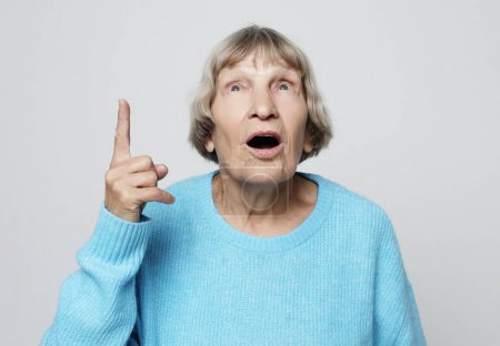 Foto de Mujer anciana sorprendida mira hacia arriba y levanta su dedo sobre fondo gris. - Imagen libre de derechos