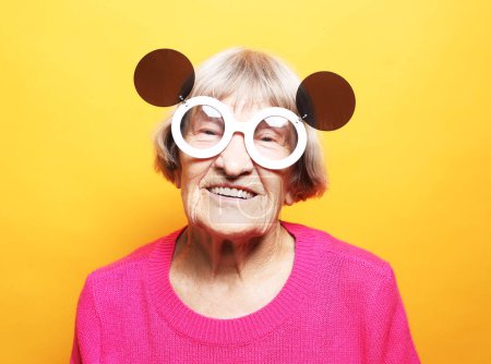 Foto de Alegre anciana vistiendo suéter rosa en grandes gafas de sol se ríe y se regocija sobre fondo amarillo - Imagen libre de derechos
