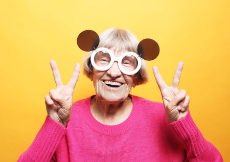 Foto de Vieja mujer feliz con gafas de sol grandes reír y mostrar la paz o la señal de victoria. Emoción y sentimientos. Retrato de la abuela expresiva sobre fondo amarillo. - Imagen libre de derechos