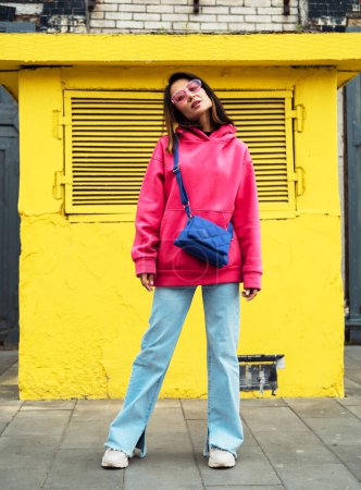 Téléchargez les photos : Belle jeune femme asiatique élégante en sweat-shirt rose et jeans posant tout en regardant la caméra - en image libre de droit
