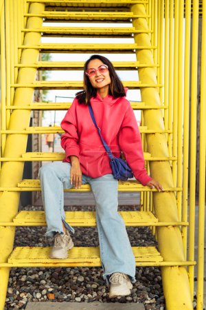 Téléchargez les photos : Jeune femme asiatique à la mode en sweat-shirt rose, lunettes de soleil et jeans est assis sur les marches des escaliers jaunes, en s'amusant. Le concept de jeunesse et de mode. - en image libre de droit
