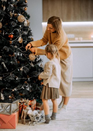 Téléchargez les photos : Famille, vacances d'hiver et concept de personnes. Joyeux jeune mère et petite fille décorant l'arbre de Noël à la maison. Amour et bonheur. - en image libre de droit