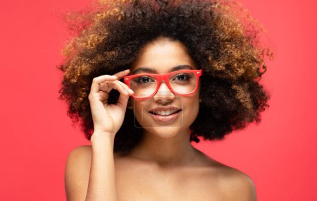 Téléchargez les photos : Portrait de belle jeune femme afro-américaine souriante. Modèle femelle avec des lunettes afro. Fond rouge. Plan studio. - en image libre de droit