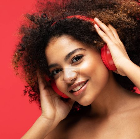 Foto de Feliz joven afroamericana mujer relajada con gran canción, tiene una amplia sonrisa, toca los auriculares, escucha música aislada sobre la pared del estudio rojo. Concepto de estilo de vida y personas. - Imagen libre de derechos