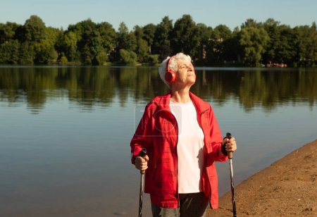 Téléchargez les photos : A pretty elderly woman with gray hair and a short haircut in a red jacket walks with trekking poles and listens to music. Summer day. The concept of a healthy lifestyle. - en image libre de droit