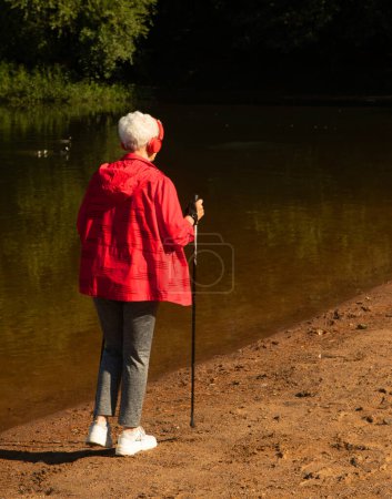 Téléchargez les photos : A pretty elderly woman with gray hair and a short haircut in a red jacket walks with trekking poles and listens to music. Summer day. View from the back. - en image libre de droit