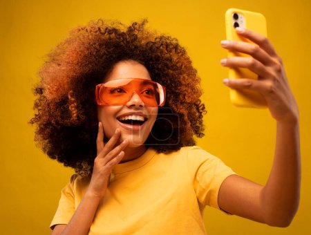 Téléchargez les photos : Jeune femme mulâtre à la peau foncée avec coiffure afro, tient un téléphone portable, passe un appel vidéo, vêtue de vêtements décontractés et de lunettes de soleil orange, isolée au-dessus du mur jaune du studio. - en image libre de droit