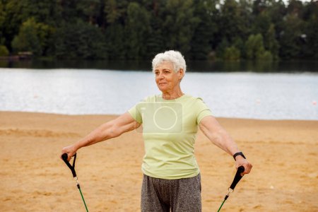 Téléchargez les photos : Old woman with short hair wear sportswear doing exercises outdoor on the beach in the morning using resistance rubber bands. Healthy lifestyle, active retired life and sporty time. - en image libre de droit
