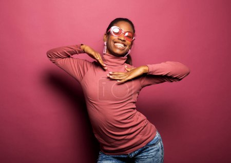 Téléchargez les photos : Photo de belle jeune femme afro porter des vêtements décontractés dansant à la recherche d'espace vide isolé fond de couleur rose. Concept de parti et de personnes. - en image libre de droit