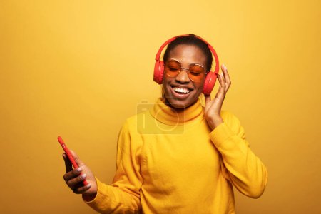 Photo for People, music, emotions concept. Delighted carefree afro american female with dances in rhythm of melody, listens loud song in headphones, holds smartphone has fun. - Royalty Free Image