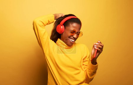 Photo for People, music, emotions concept. Delighted carefree afro american female with dances in rhythm of melody, listens loud song in headphones, holds smartphone has fun. - Royalty Free Image